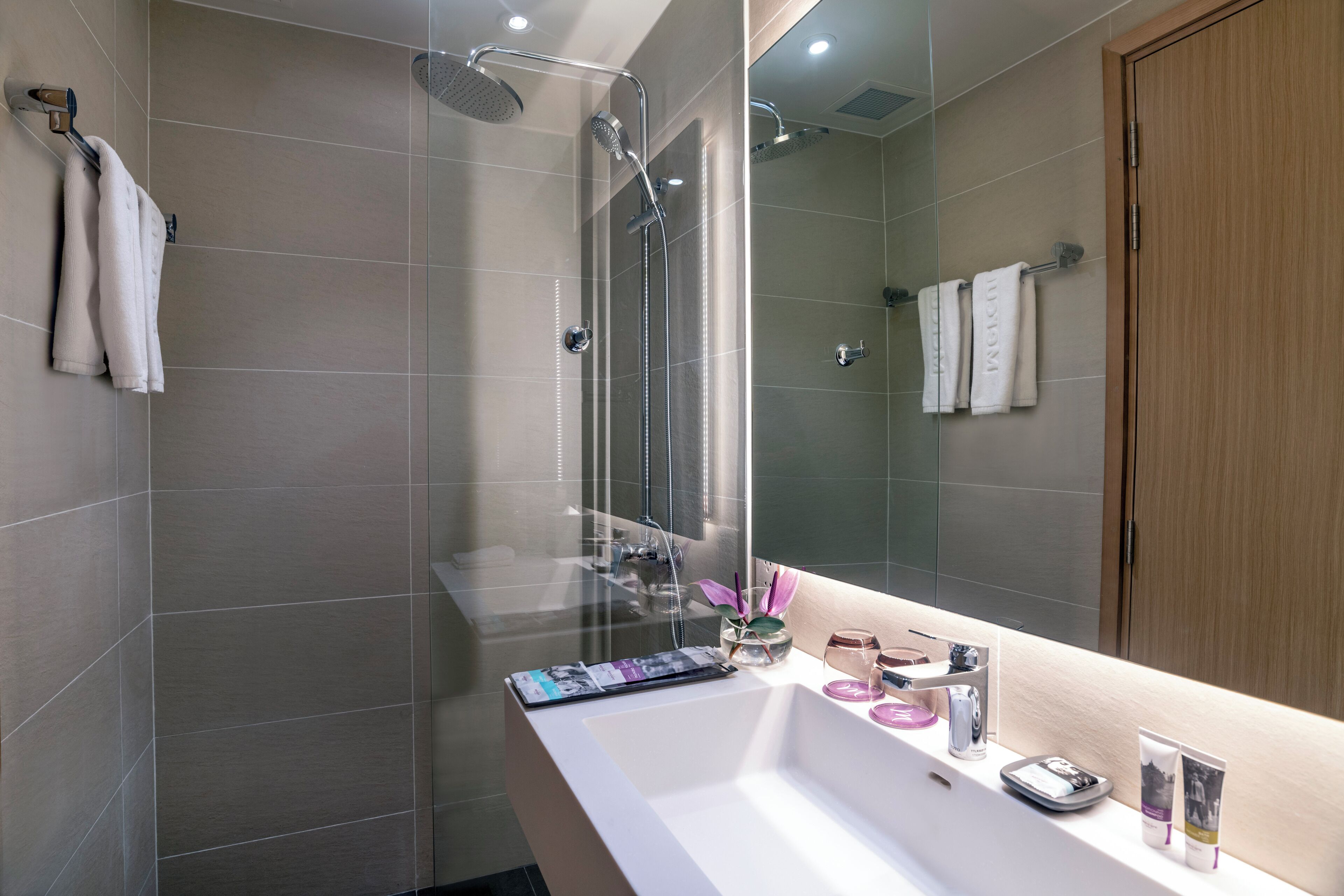 Une salle de bain moderne avec une douche à parois de verre, un lavabo blanc agrémenté d’articles de toilette et une porte en bois.