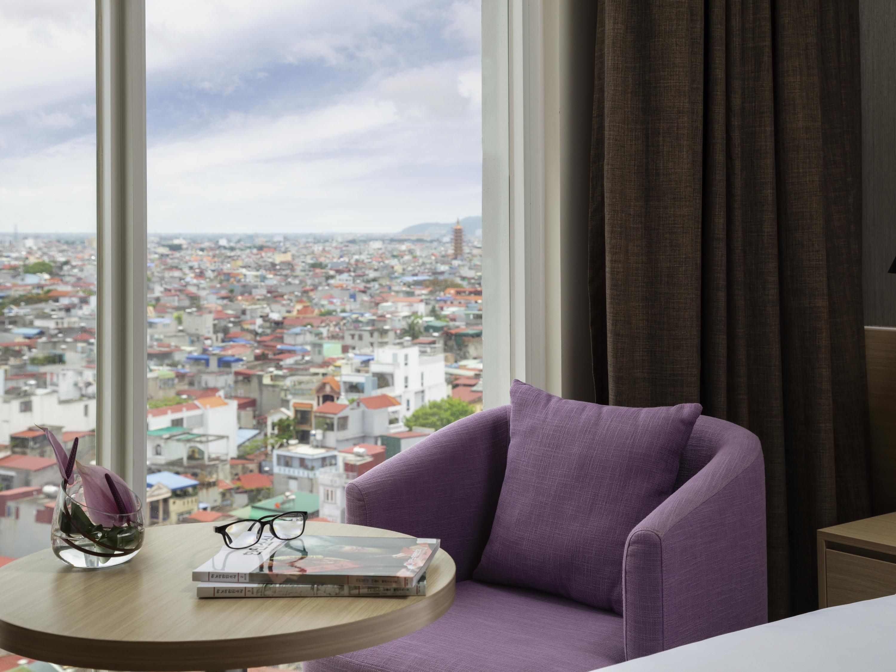 A room with a view of a cityscape through large windows, a purple armchair, a small round table with a book and glasses, and a vase with a plant.
