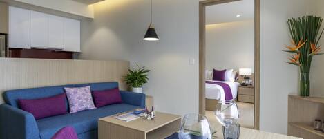 A modern hotel room with a blue sofa, a dining table set with cutlery and glasses, a mirror, and a potted plant.