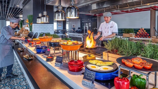 Een moderne keuken met koks die eten bereiden, een buffet met diverse gerechten en een presentatie van vers fruit.