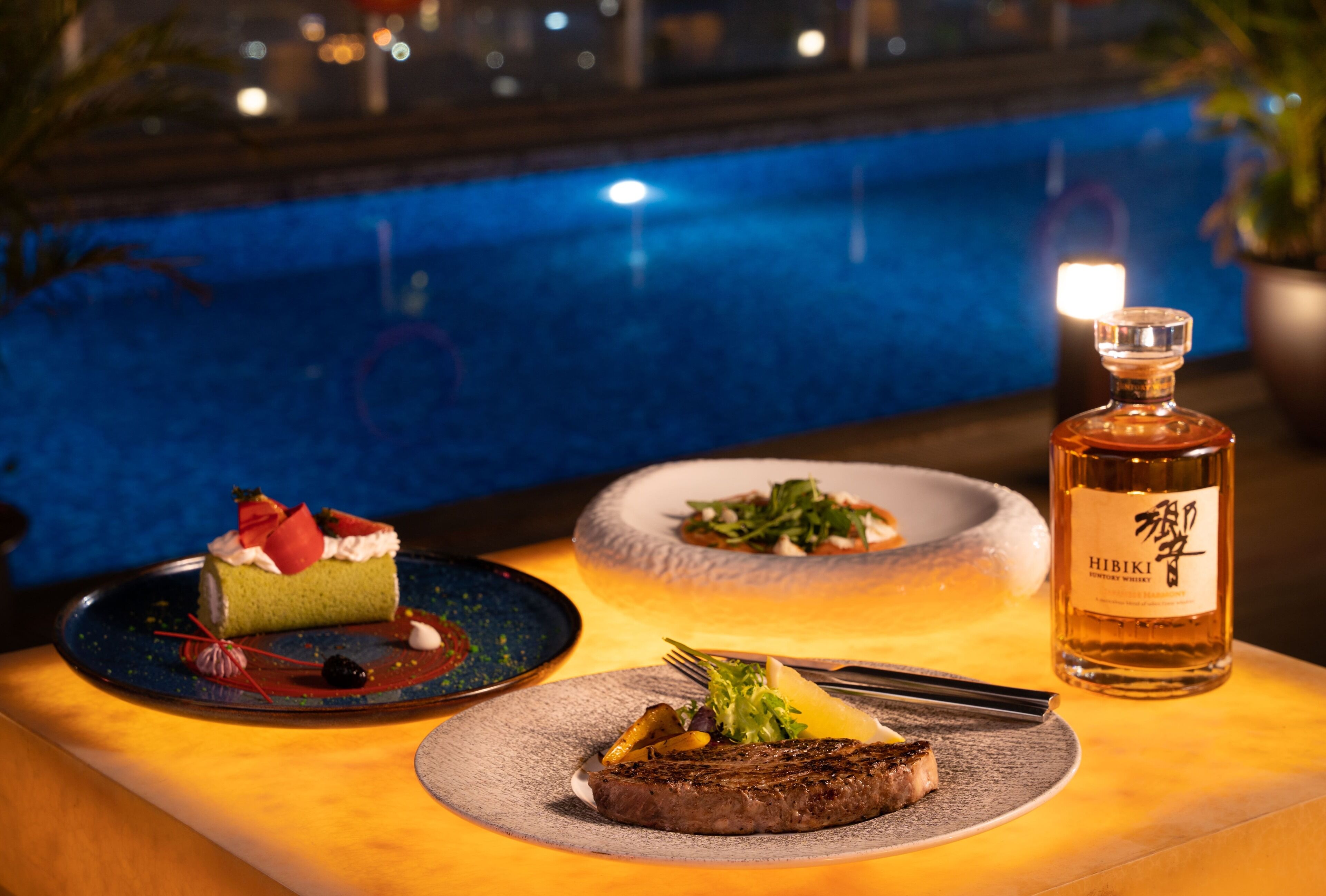 Una mesa de comedor con un bistec, un postre y una botella de whisky Hibiki.