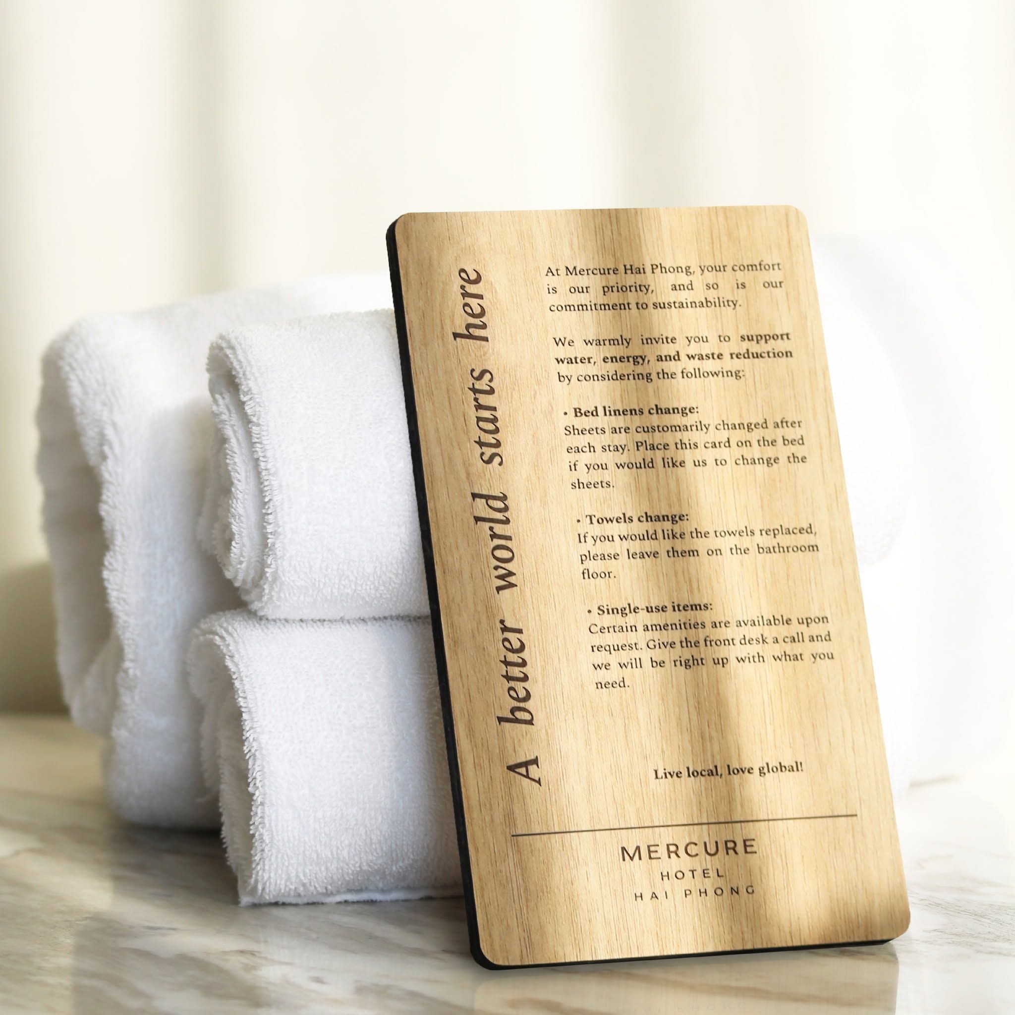 A wooden card with text about sustainability practices at Mercure Hai Phong hotel, placed next to folded white towels.