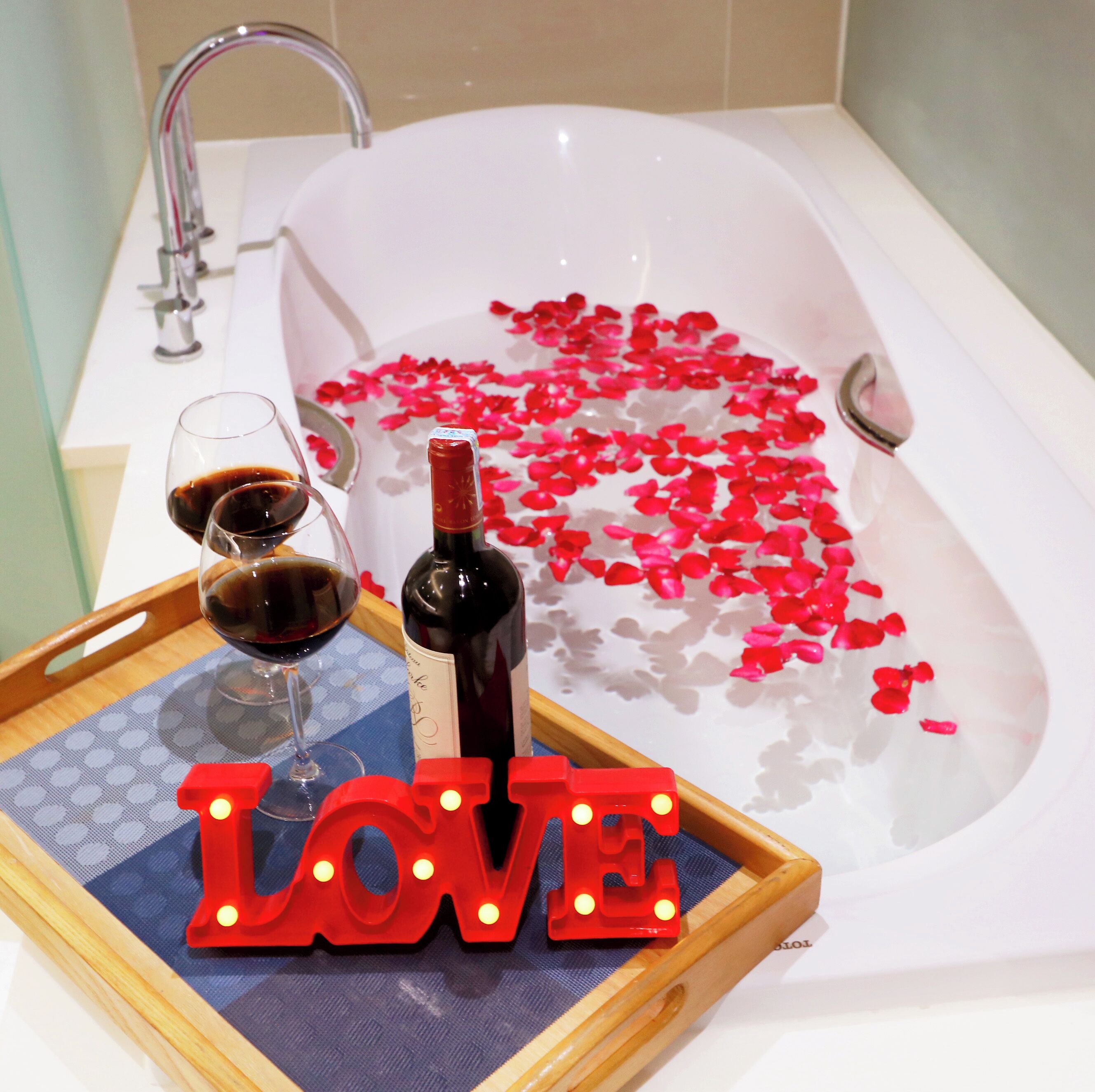 A bathtub filled with rose petals and a bottle of wine, with glasses and a tray with the word "LOVE" on it.