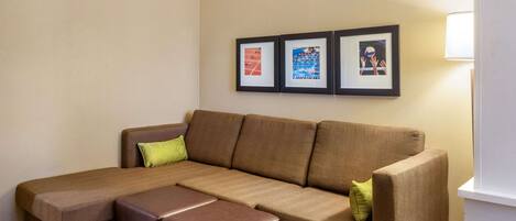 A brown sectional sofa with a matching ottoman, a green pillow, and a framed picture of a tennis court on the wall.
