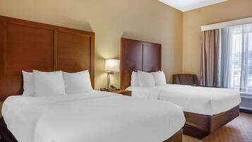 Habitación de hotel con dos camas, cabecera de madera, mesita de noche con lámpara y ventana grande con cortinas.