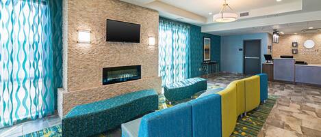 A hotel lobby with a stone wall, a flat-screen TV, a fireplace, and a seating area with blue and yellow sofas.
