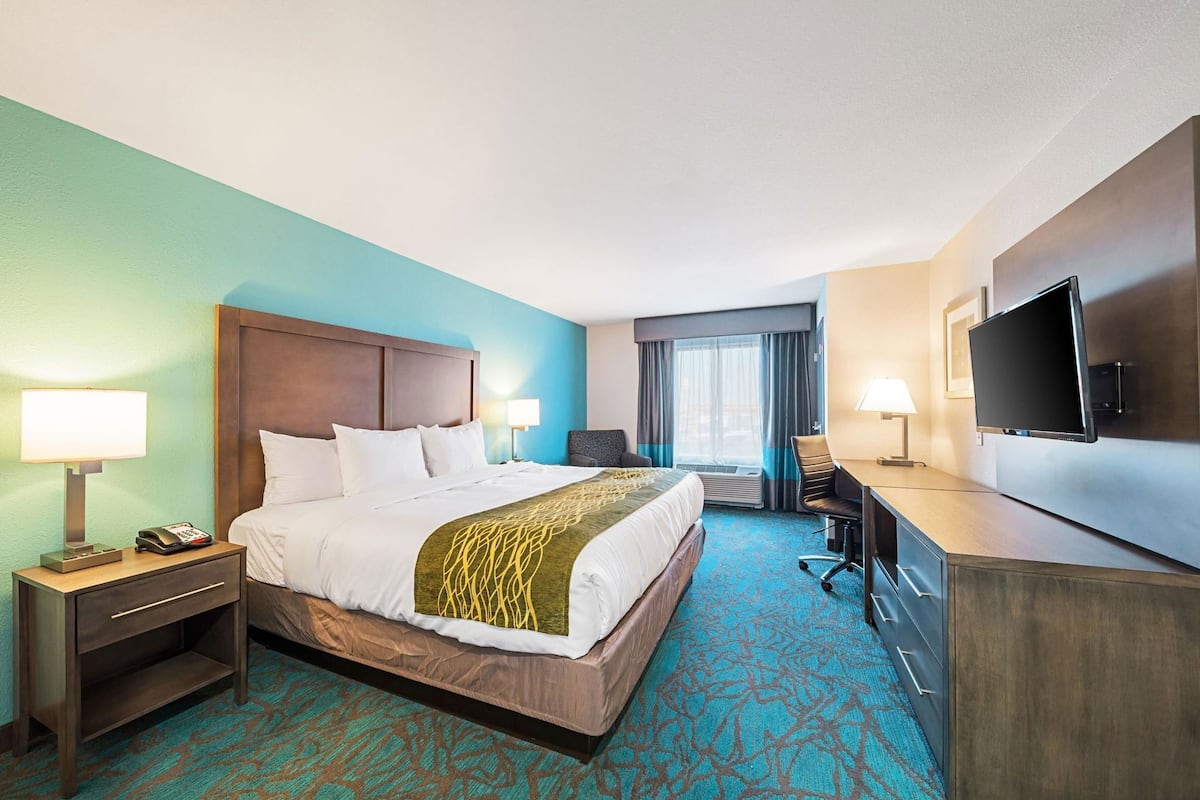 Ein Hotelzimmer mit einem großen Bett, einem Schreibtisch mit Flachbildfernseher, einem Sessel, einer Schreibtischlampe, einem Telefon und einem Nachttisch.