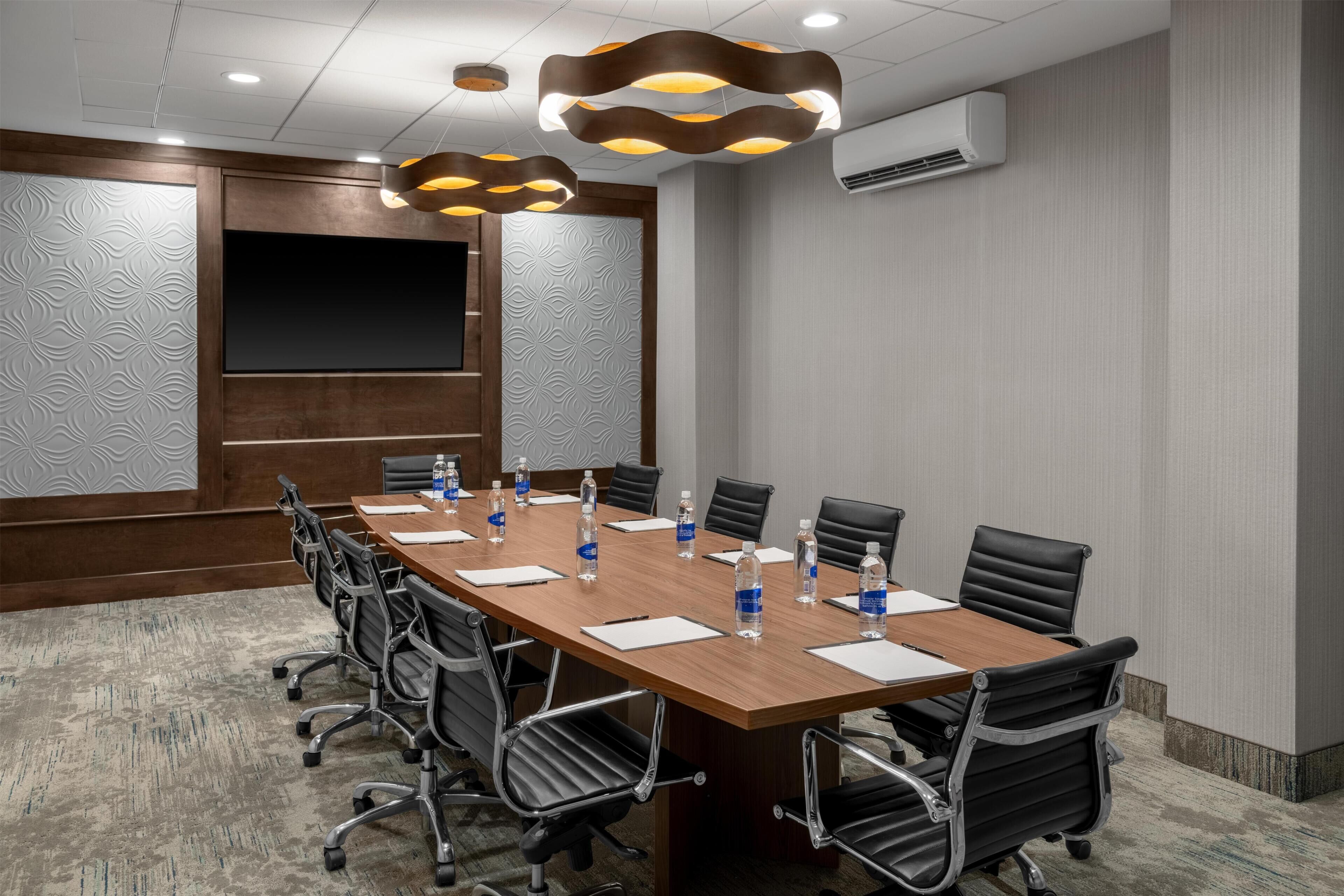 Uma sala de conferências com uma mesa comprida, cadeiras, garrafas de água e uma tela de projetor.