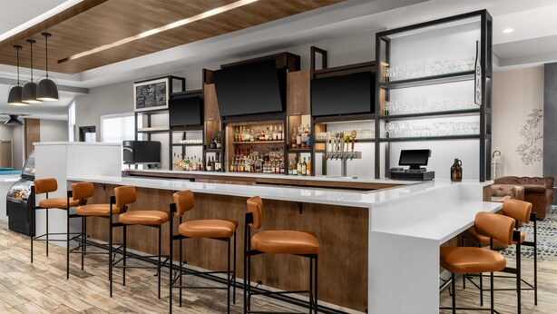 A modern bar area with a long counter, multiple stools, and a variety of bottles and glasses. - Hilton Garden Inn Topeka (Topeka)