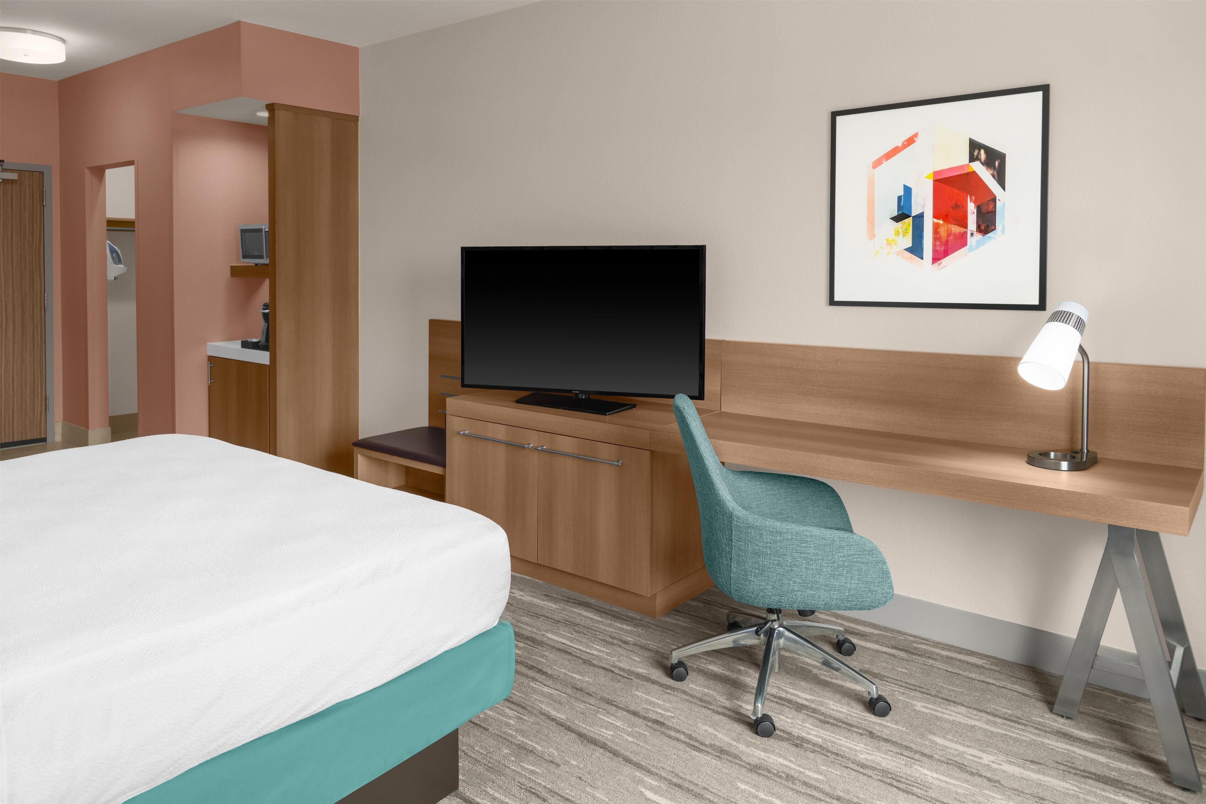 Habitación de hotel con cama, escritorio con computadora, televisor, lámpara de escritorio y un cuadro en la pared.