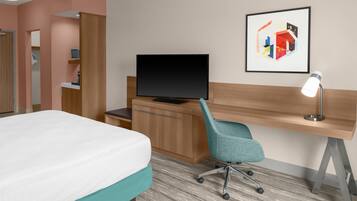 Habitación de hotel con cama, escritorio con computadora, televisor, lámpara de escritorio y un cuadro en la pared.