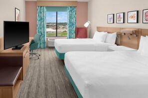 Ein Hotelzimmer mit zwei Betten, einem Schreibtisch mit Fernseher, einem Sessel und einem Fenster mit Vorhängen.
