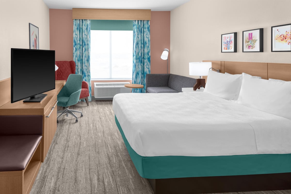A hotel room with a bed, a desk with a TV, a chair, a lamp, and a window with curtains.