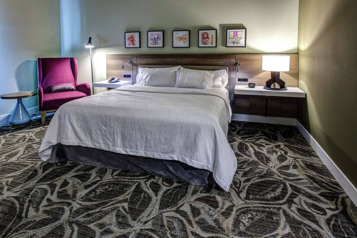 A hotel room with a bed, a chair, a nightstand, and a wall with framed pictures.