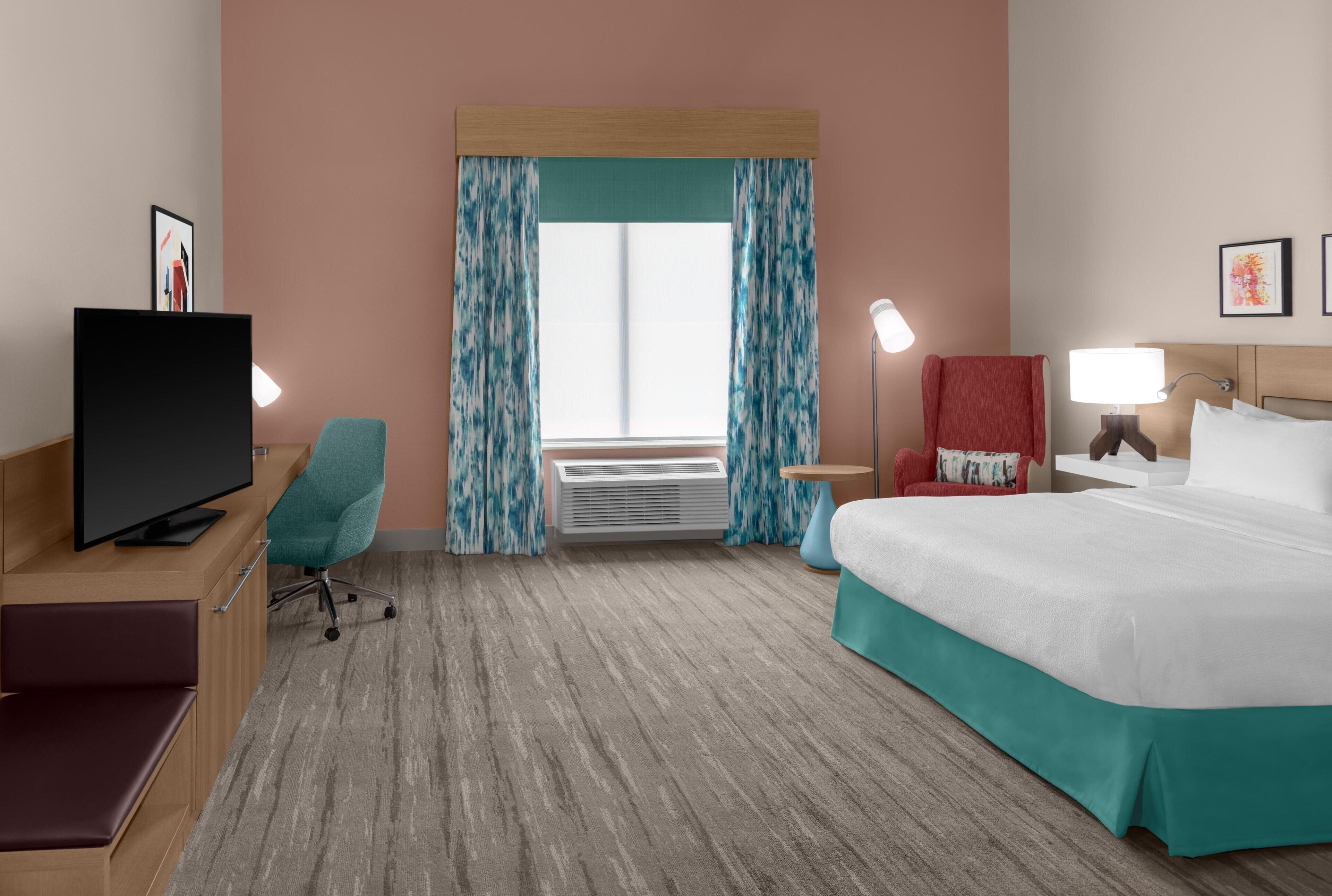 Une chambre d’hôtel avec un lit, un bureau avec une télévision, une chaise, une lampe et une fenêtre avec des rideaux.