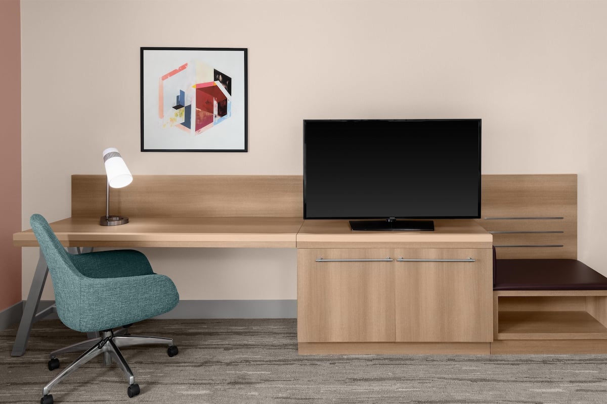 A modern office desk with a television, a teal office chair, a desk lamp, and a framed abstract artwork on the wall.