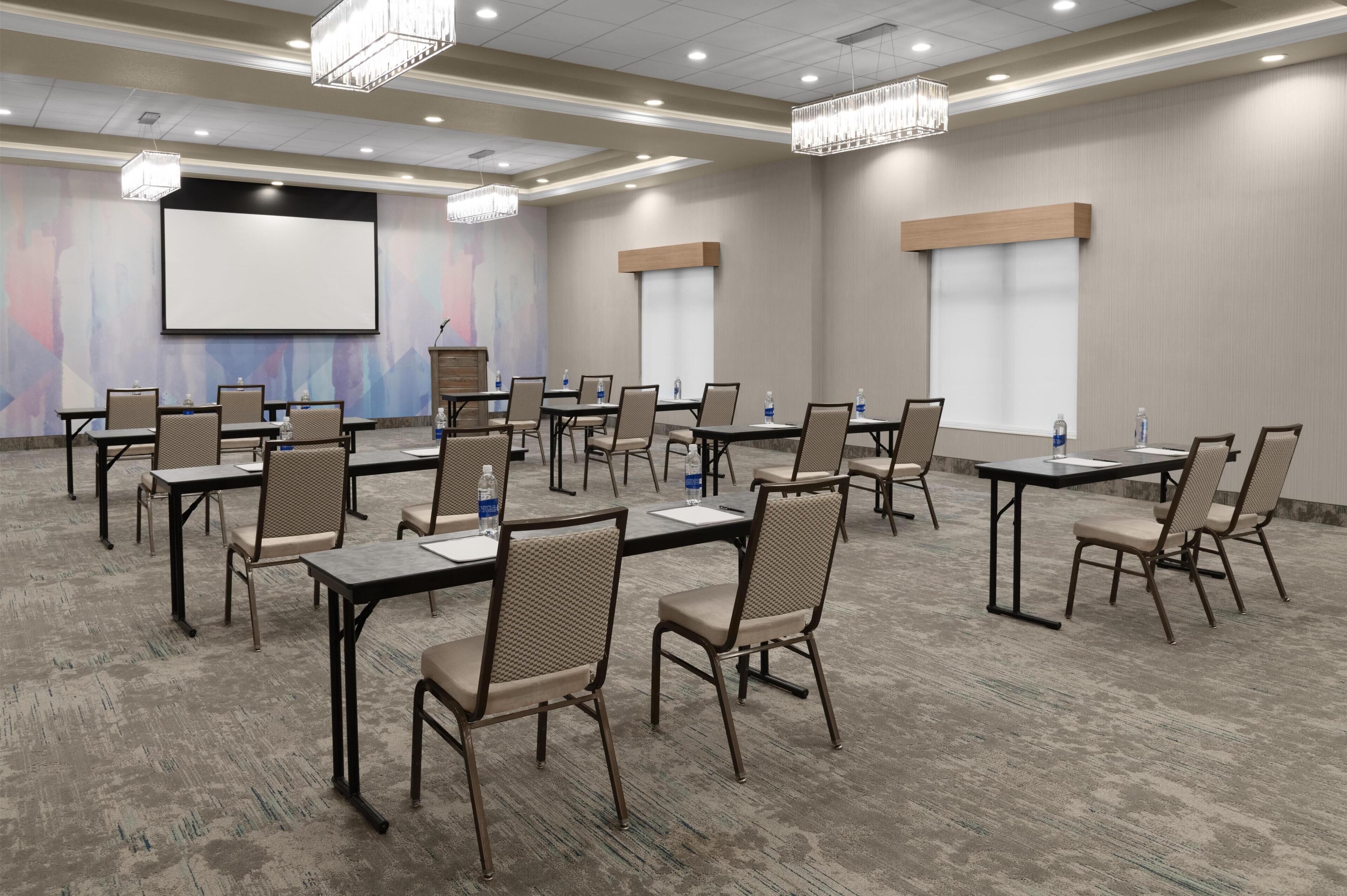 Une salle de conférence équipée de tables et de chaises, d’un écran de projection et d’un grand mur vierge.