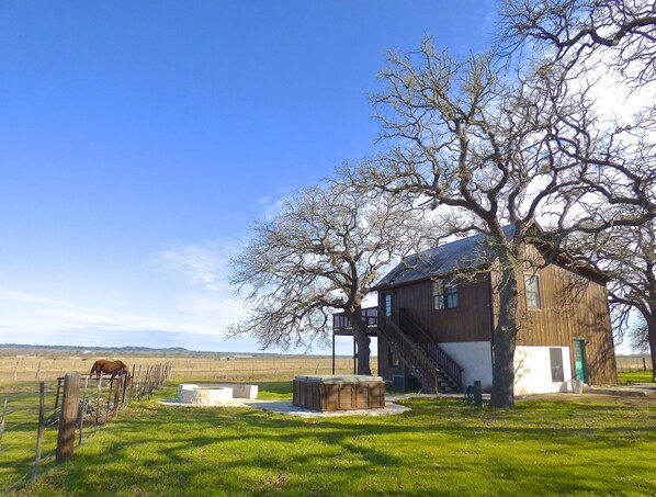 A rustic wooden building with a staircase leading to a loft area, surrounded by a fenced pasture with a horse.