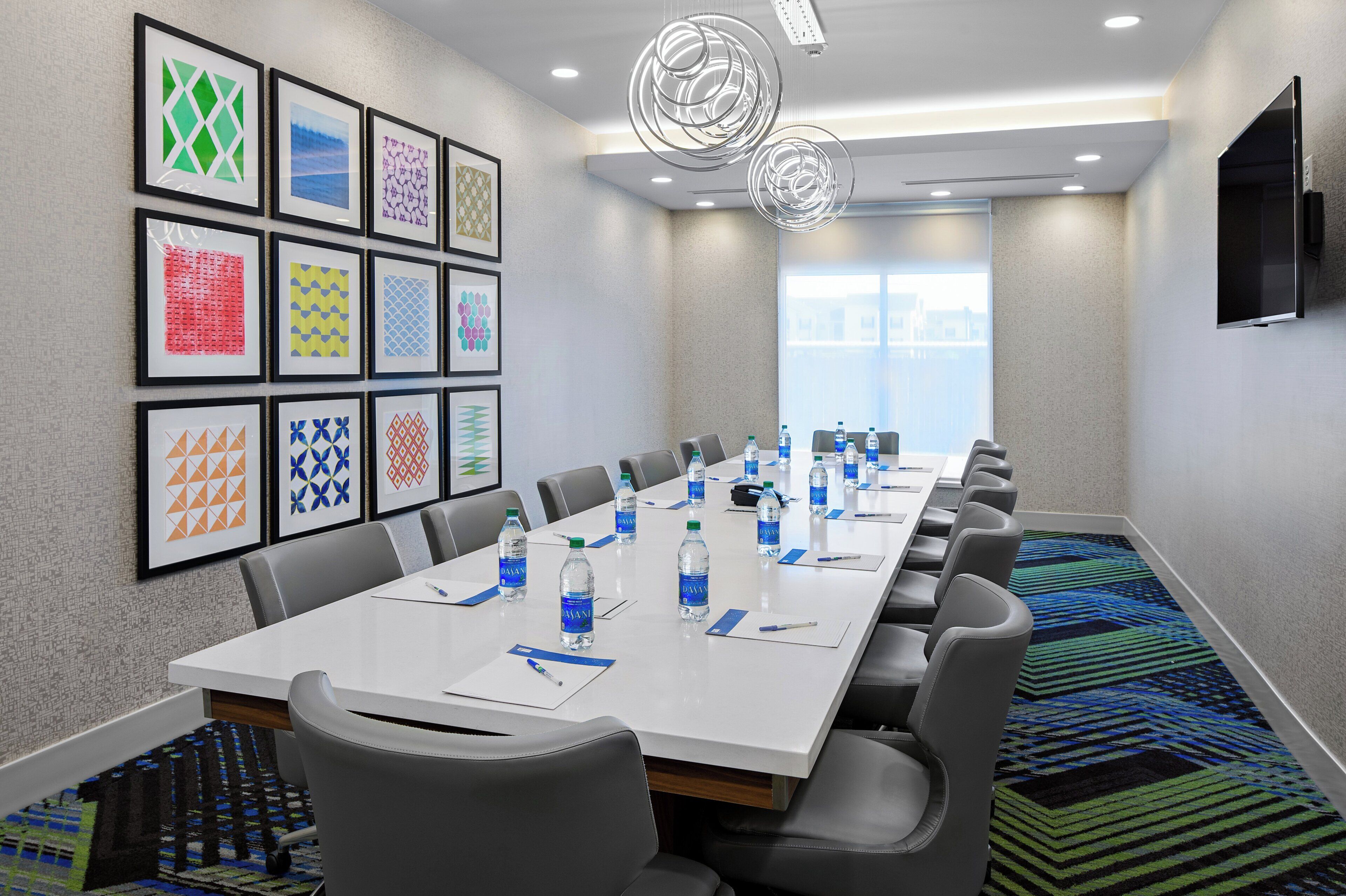 A conference room with a long table, chairs, framed artwork on the wall, water bottles, and a TV on the wall.