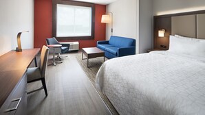 A hotel room with a large bed, a desk, a chair, a sofa, a small table, a lamp, and a window.