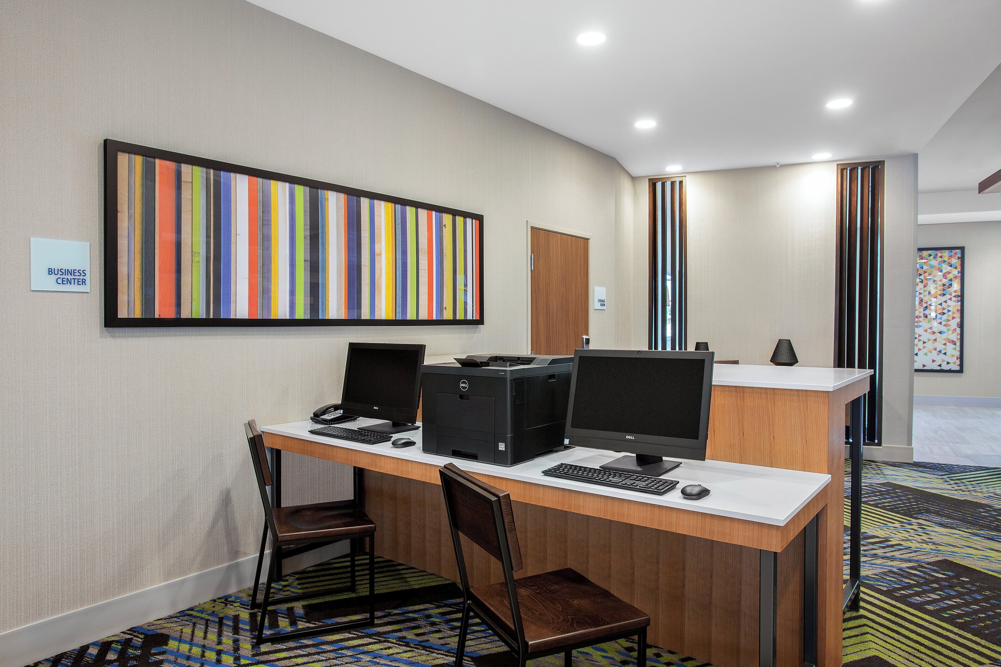 A hotel business center with a desk, computer, printer, and chairs.