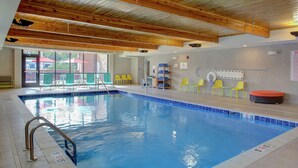 Una piscina interior con techo de madera, paredes de vidrio transparente y sillas amarillas alrededor del borde.