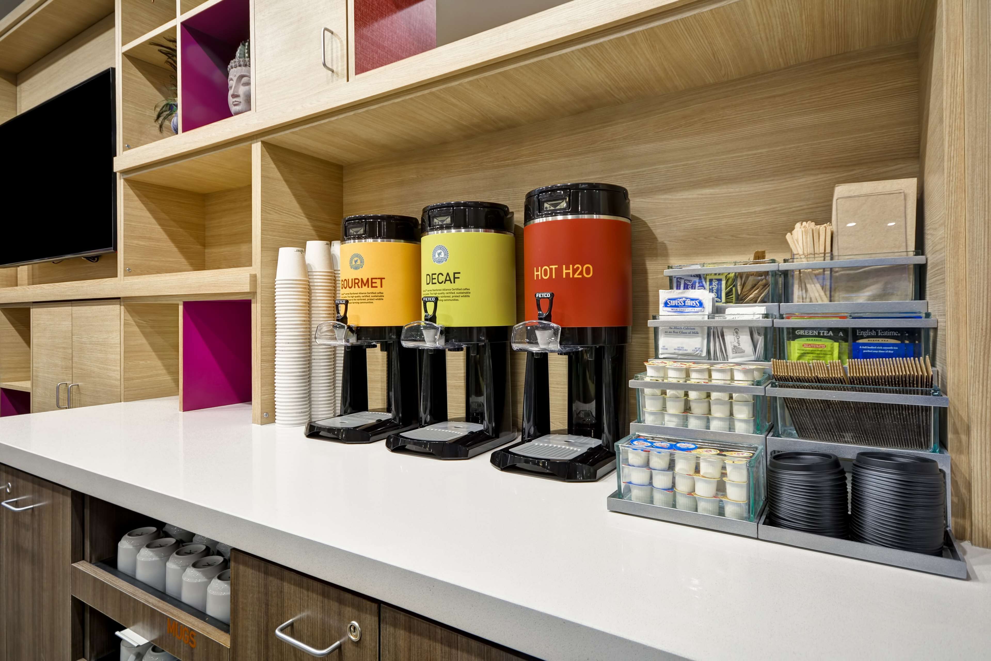 A coffee station with labeled dispensers for Gourmet, Decaf, and Hot H2O beverages, and a tray of black disposable cups.