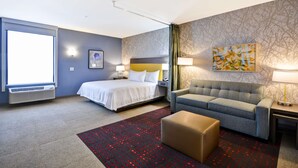 Habitación de hotel con cama, sofá, mesita, lámpara, un cuadro y ventana.