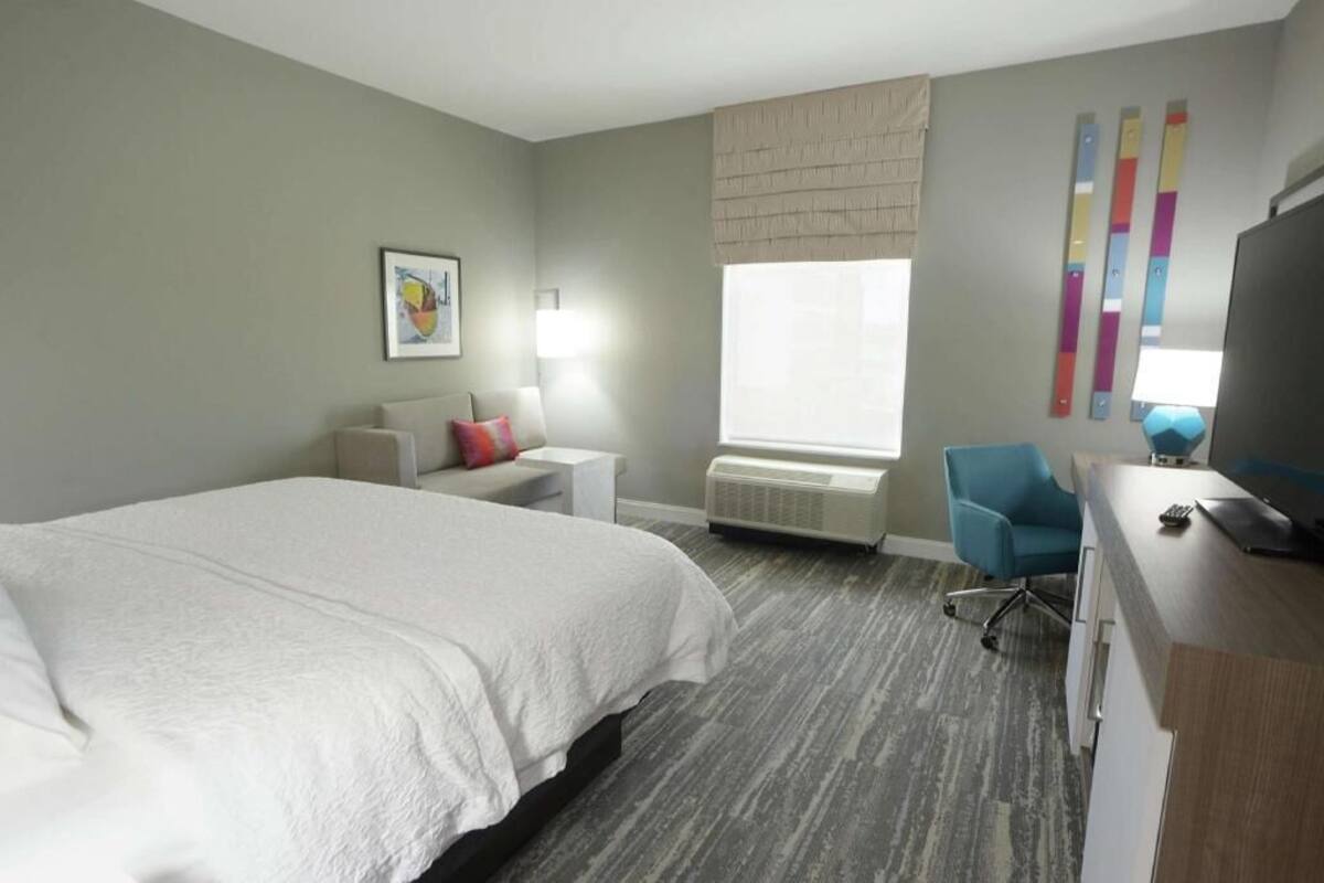 Hotelkamer met een groot bed, een bureau, een stoel, een lamp, een raam met zonwering en een wandairconditioner.