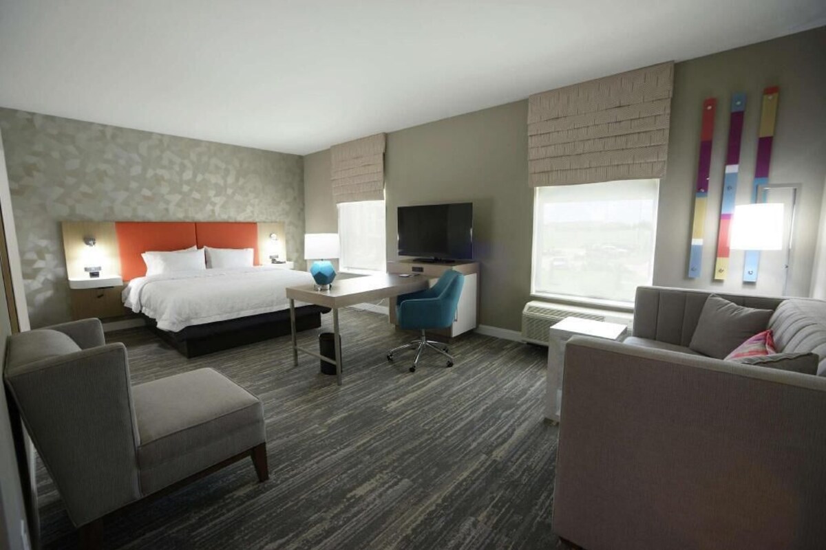 Quarto de hotel com cama, escrivaninha com cadeira, televisão, sofá e janela com persianas.