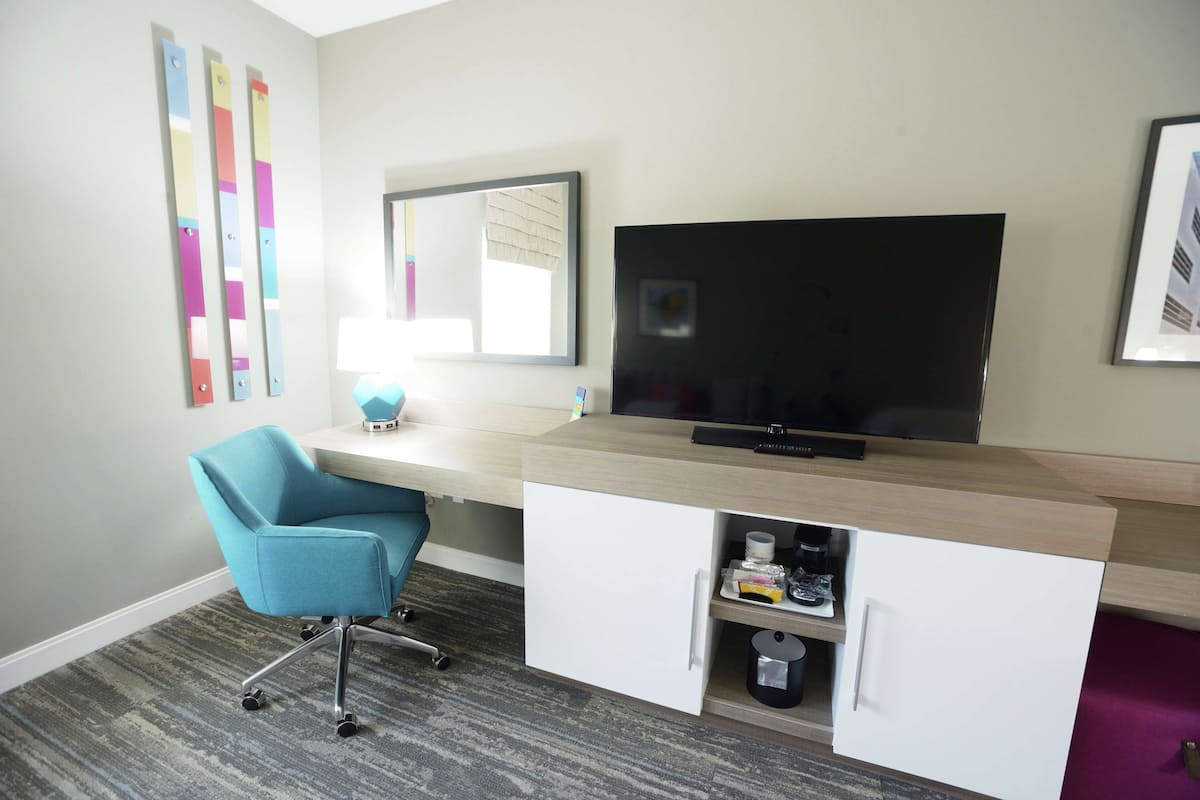 Een moderne hotelkamer met een lichtkleurig bureau, een blauwe bureaustoel, een flatscreen-televisie, een klein kastje met een koffiezetapparaat en een spiegel aan de muur.