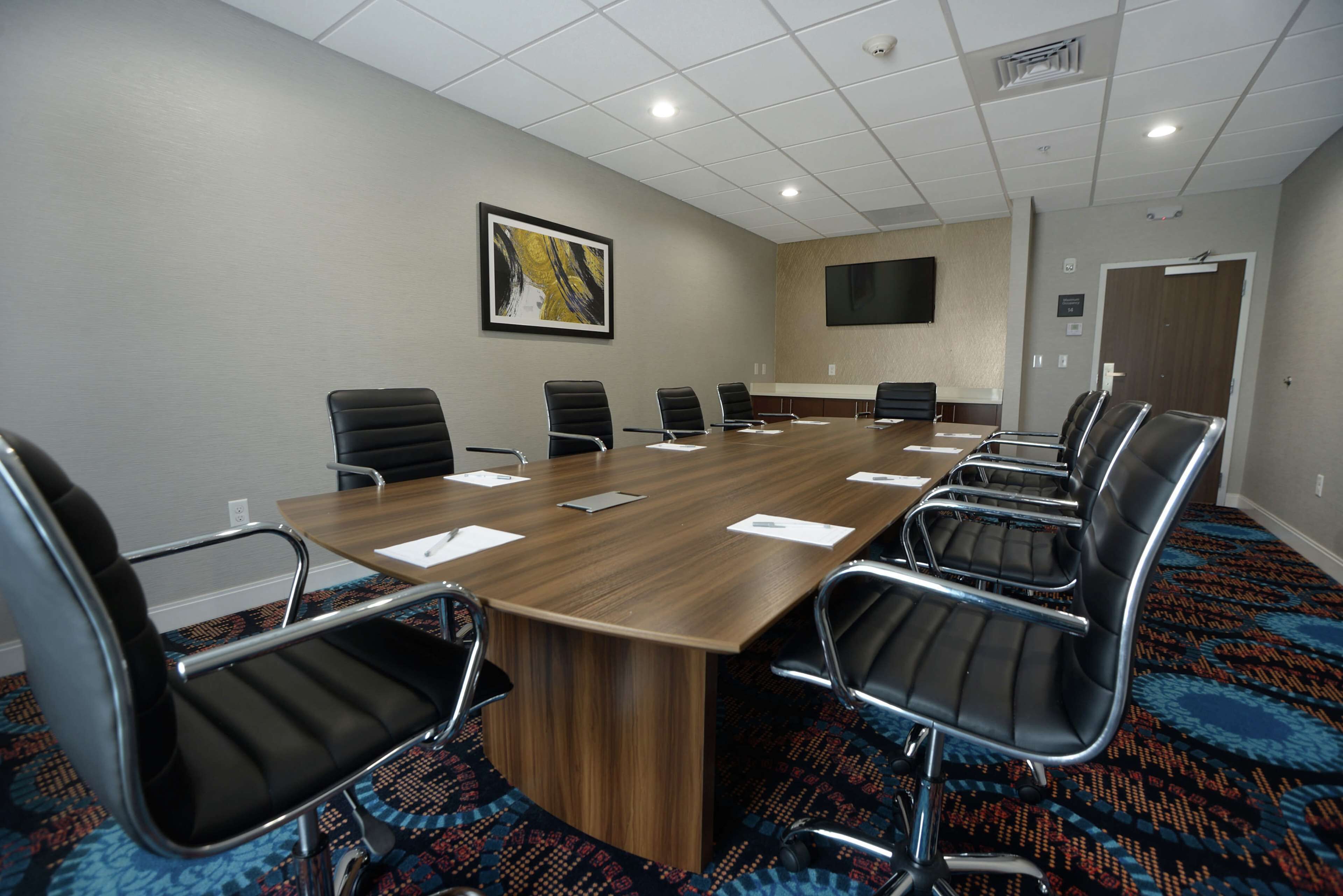 A conference room with a long wooden table, black chairs, a mounted flat-screen TV, and a patterned carpet.