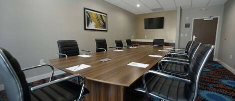 A conference room with a long wooden table, black chairs, a mounted flat-screen TV, and a patterned carpet.