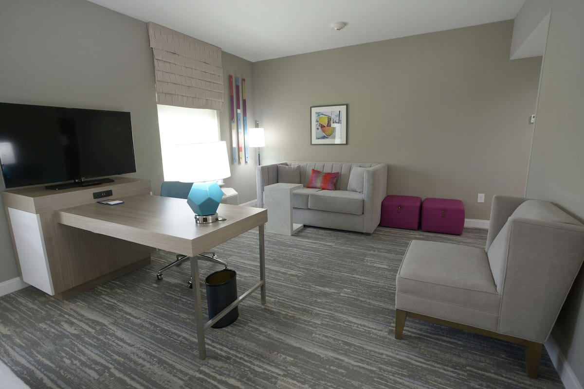 Een moderne woonkamer met een flatscreen-tv, een bureau met een lamp, een grijze bank en een roze koffer.