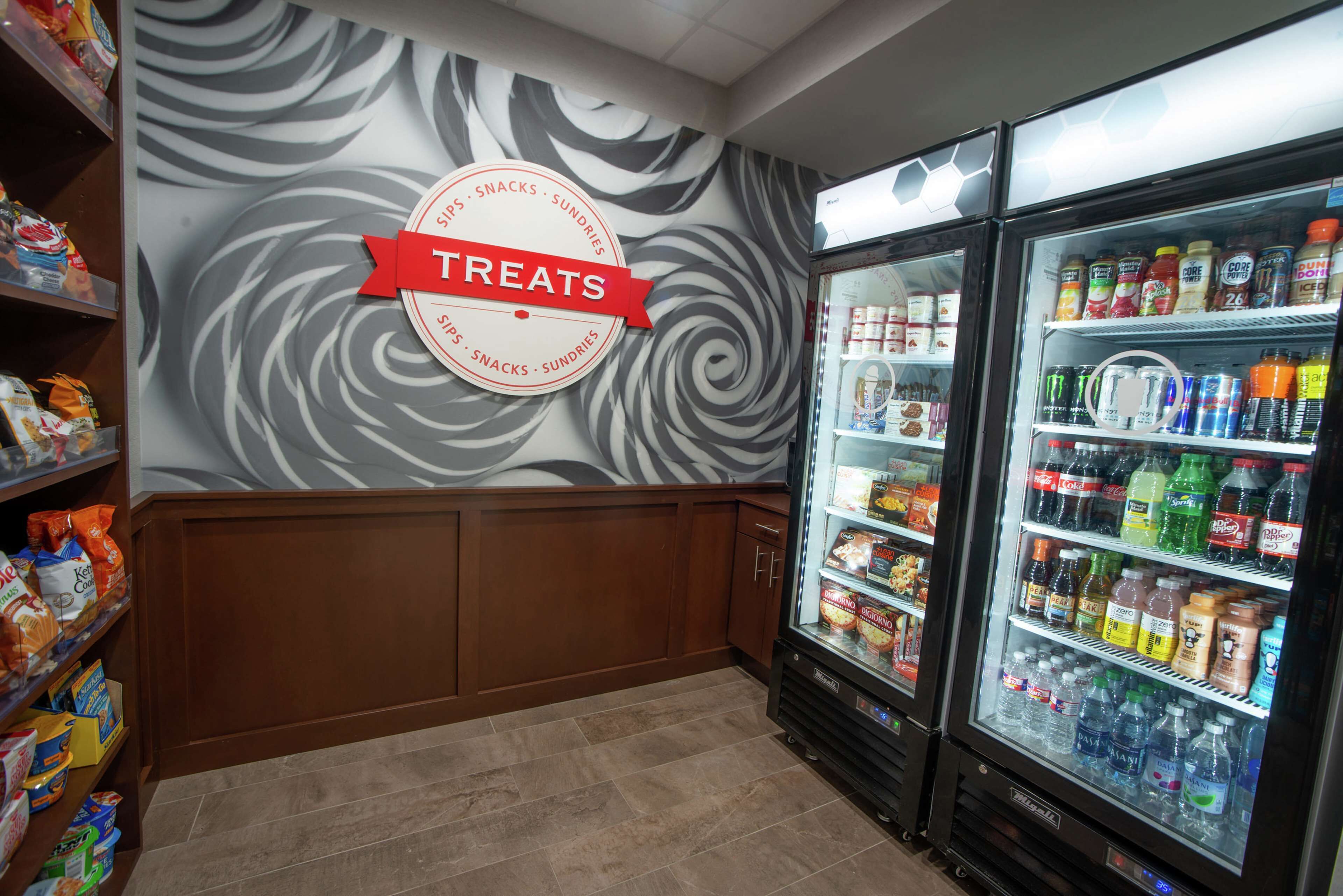 A grocery store with a 'Treats' sign on the wall, refrigerated beverage cases, and shelves stocked with snacks and groceries.