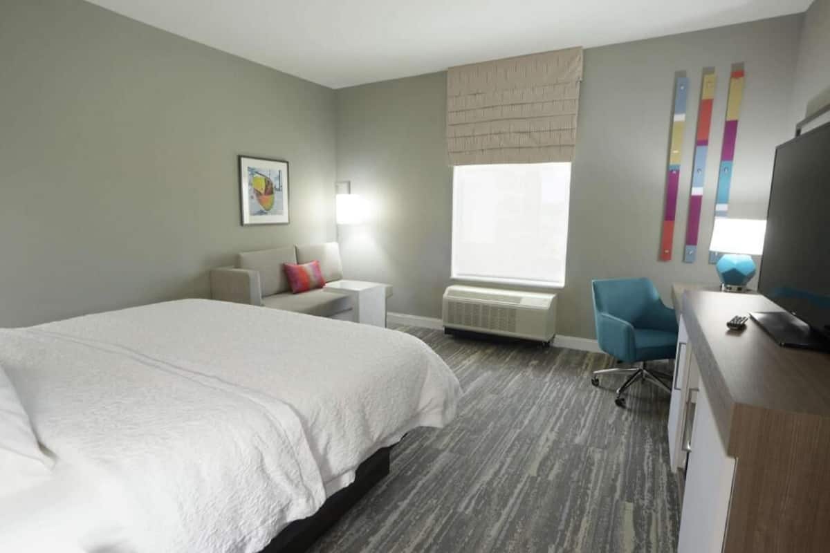 Hotelkamer met een groot bed, een bureau, een stoel, een lamp, een raam met zonwering en een wandairconditioner.