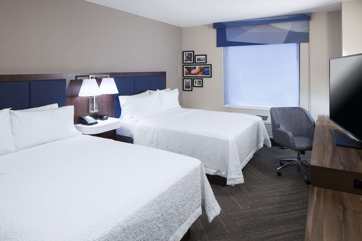 A hotel room with two beds, a desk with a lamp, a TV, and a window with a view.