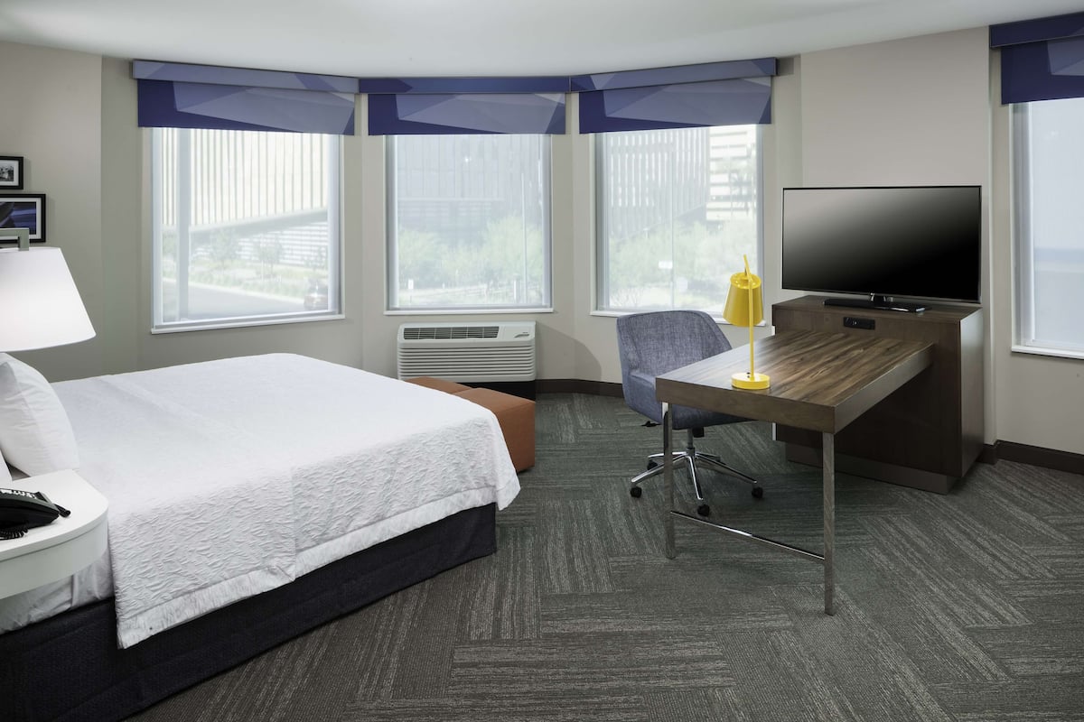 A hotel room with a bed, desk, chair, TV, and large windows.