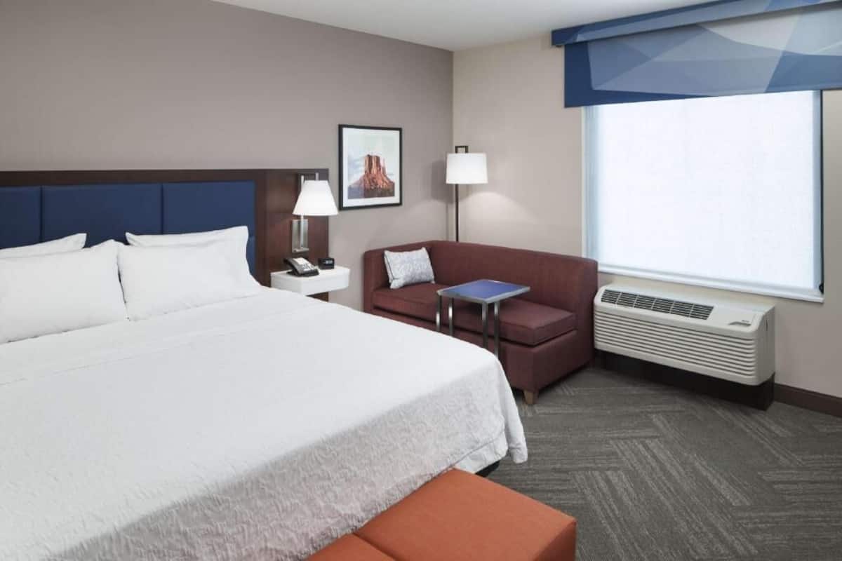 A hotel room with a large bed, a desk with a lamp, a chair, a small table, and a window with an air conditioner.