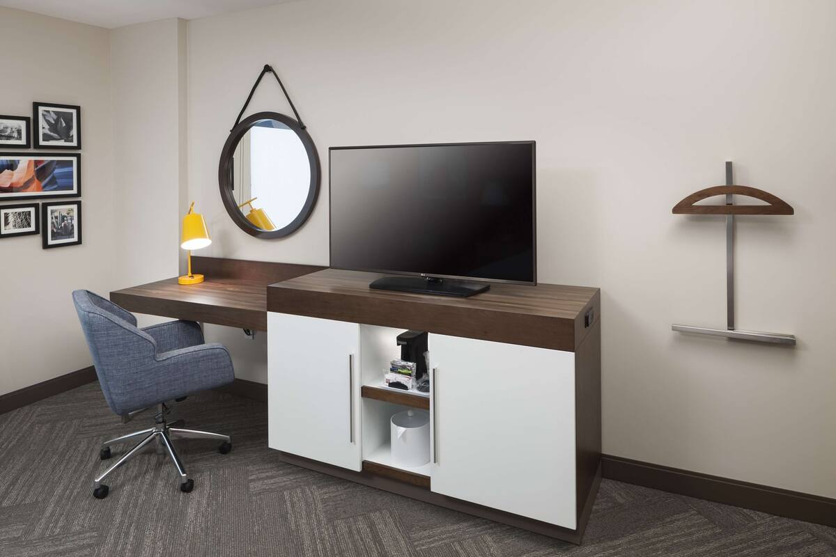 A hotel room with a desk, chair, television, mirror, and wall-mounted shelf.