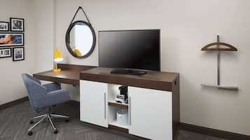 A hotel room with a desk, chair, television, mirror, and wall-mounted shelf.