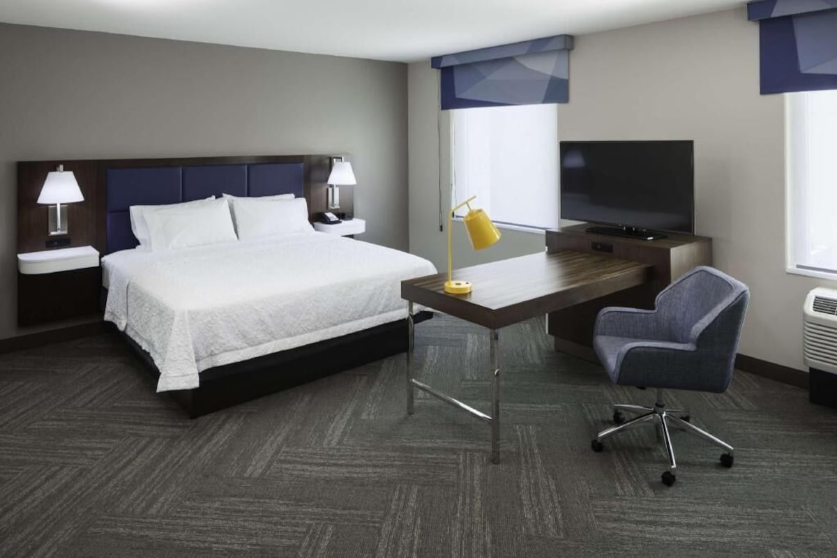 A hotel room with a bed, desk, chair, TV, lamp, and a window.
