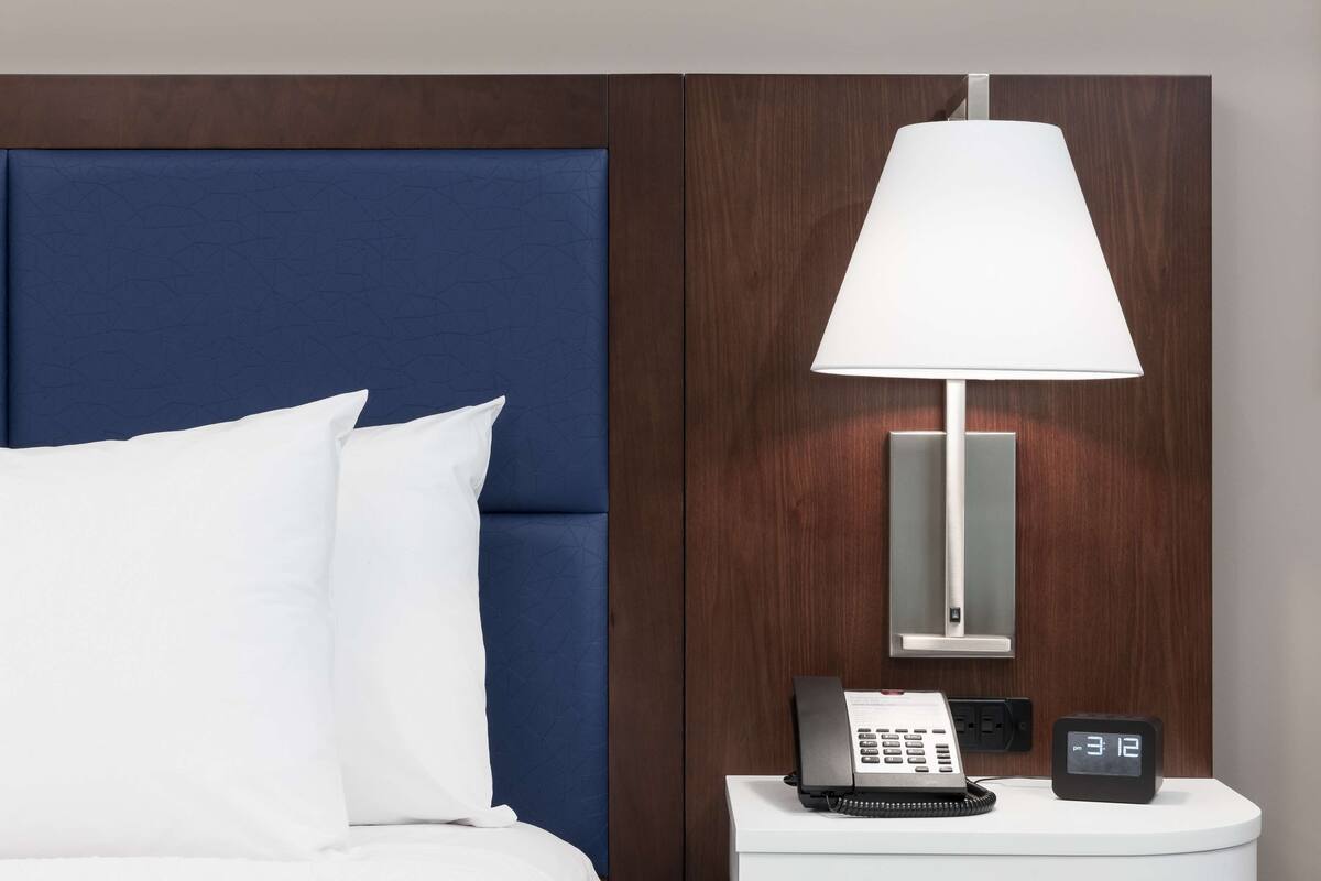 A hotel room with a blue headboard, a white bed, a bedside table with a lamp, a phone, and a digital clock.