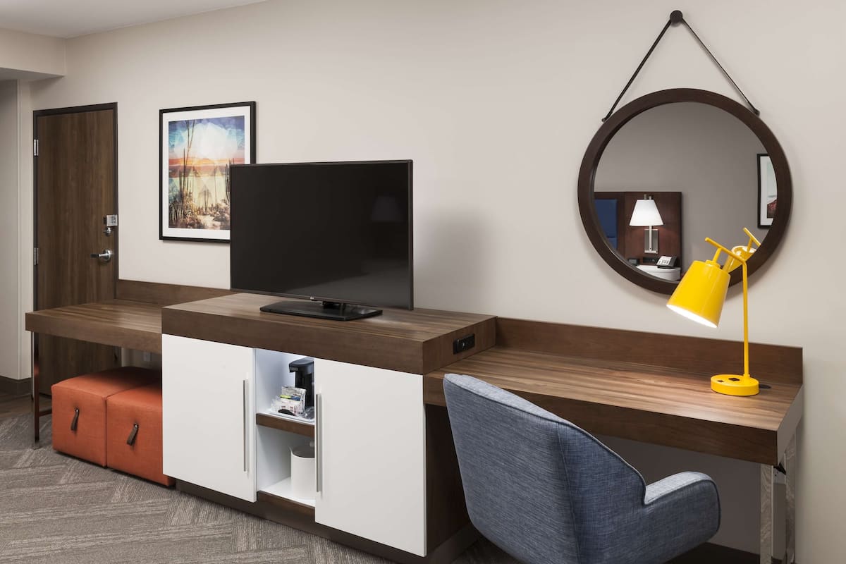 A modern hotel room with a wooden desk, a flat-screen TV, a small cabinet, a round mirror, and a yellow desk lamp.
