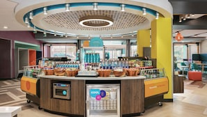A modern, well-lit indoor space with a central counter displaying various products, a digital kiosk, and a refrigerated display.