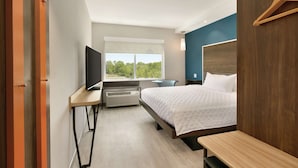 A modern hotel room with a bed, bedside table, TV, and a window with a view of greenery.