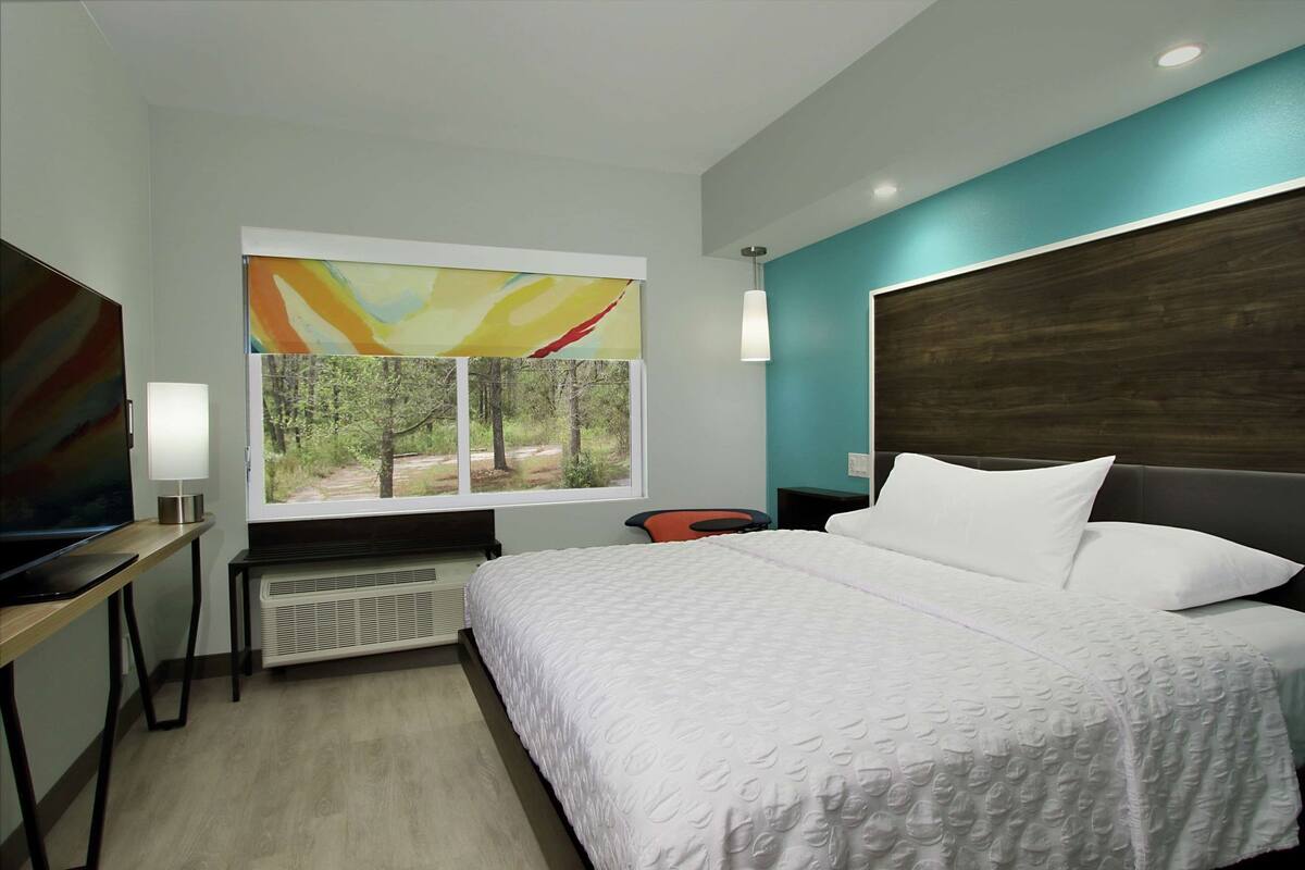 A hotel room with a bed, a desk with a lamp, a window with a view of trees, and a painting on the wall.