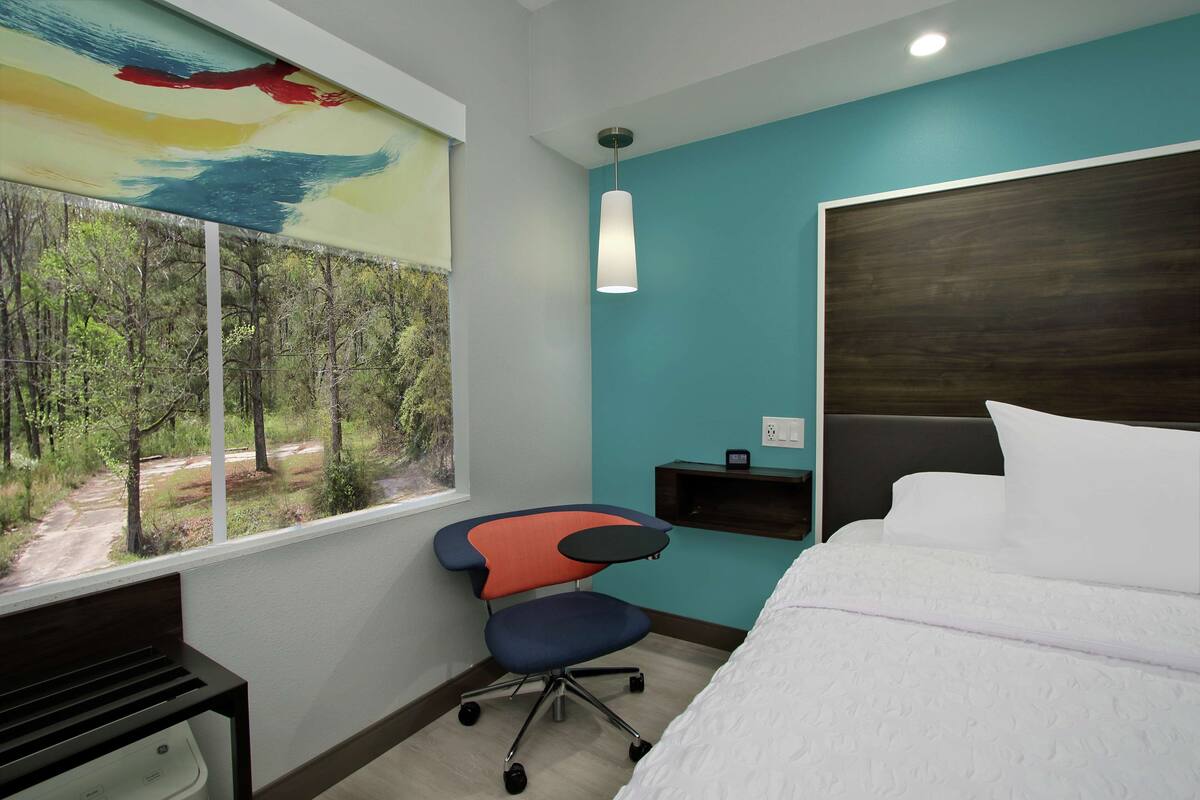 A hotel room with a bed, a desk, a chair, a window with a view of a forest, and a wall-mounted shelf.