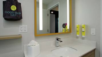 Un baño con un lavamanos blanco, un espejo, un dispensador de toallas y una bolsa de tela con la inscripción 'Good Hair Day' colgada en la pared.