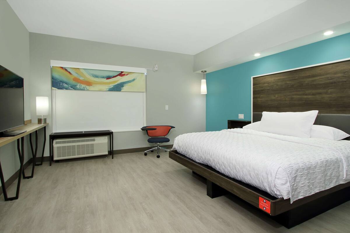 A modern hotel room with a large bed, a desk with a chair, a TV, and a painting on the wall.